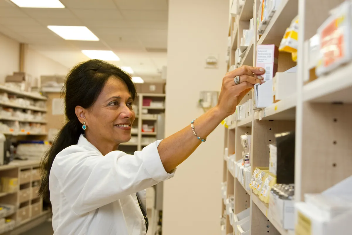 Farmacêutica de jaleco branco sorrindo enquanto pega uma caixa de medicamento na prateleira da farmácia.
